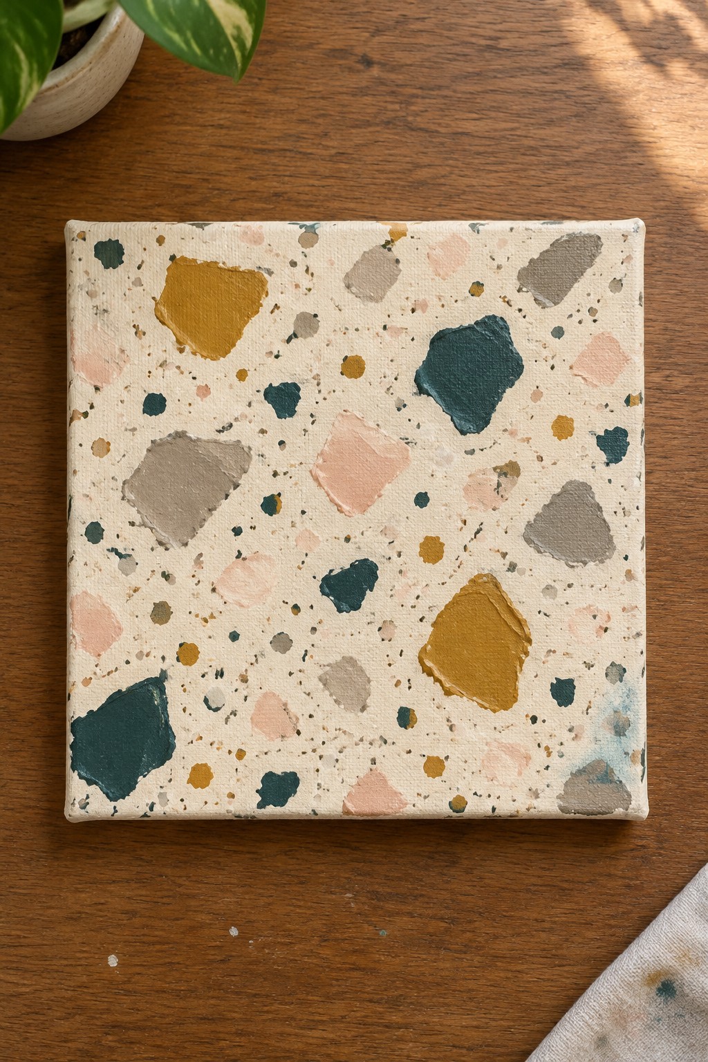 Colorful terrazzo-style abstract painting on square canvas atop wooden table with plant.