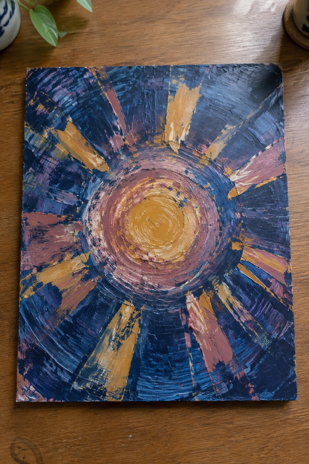Square abstract painting showing a textured yellow-orange center with radiating strokes in dark blue, purple, pink, and orange on a wooden table.