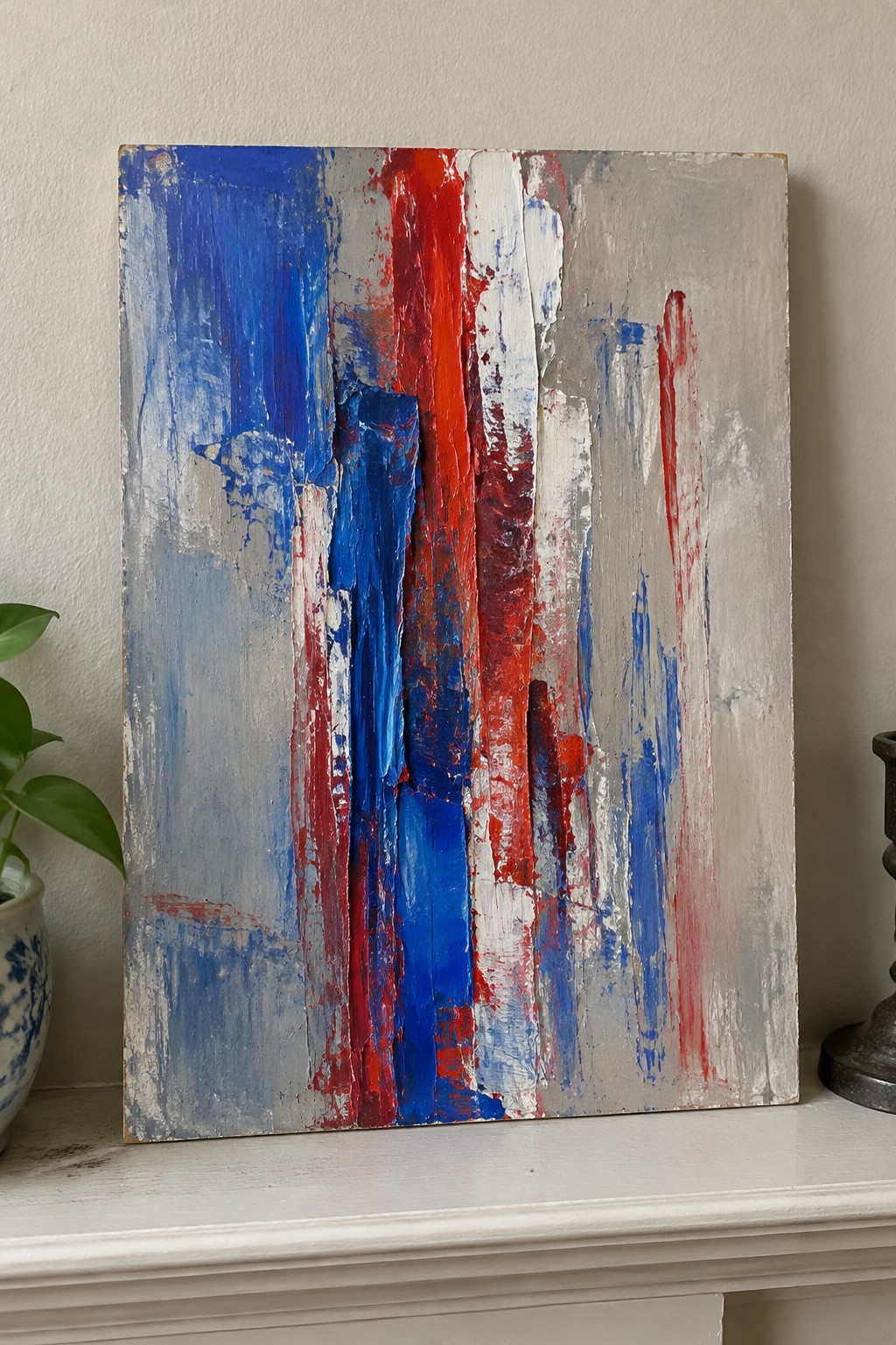 Abstract painting with thick vertical blue red white gray brushstrokes on white mantel.