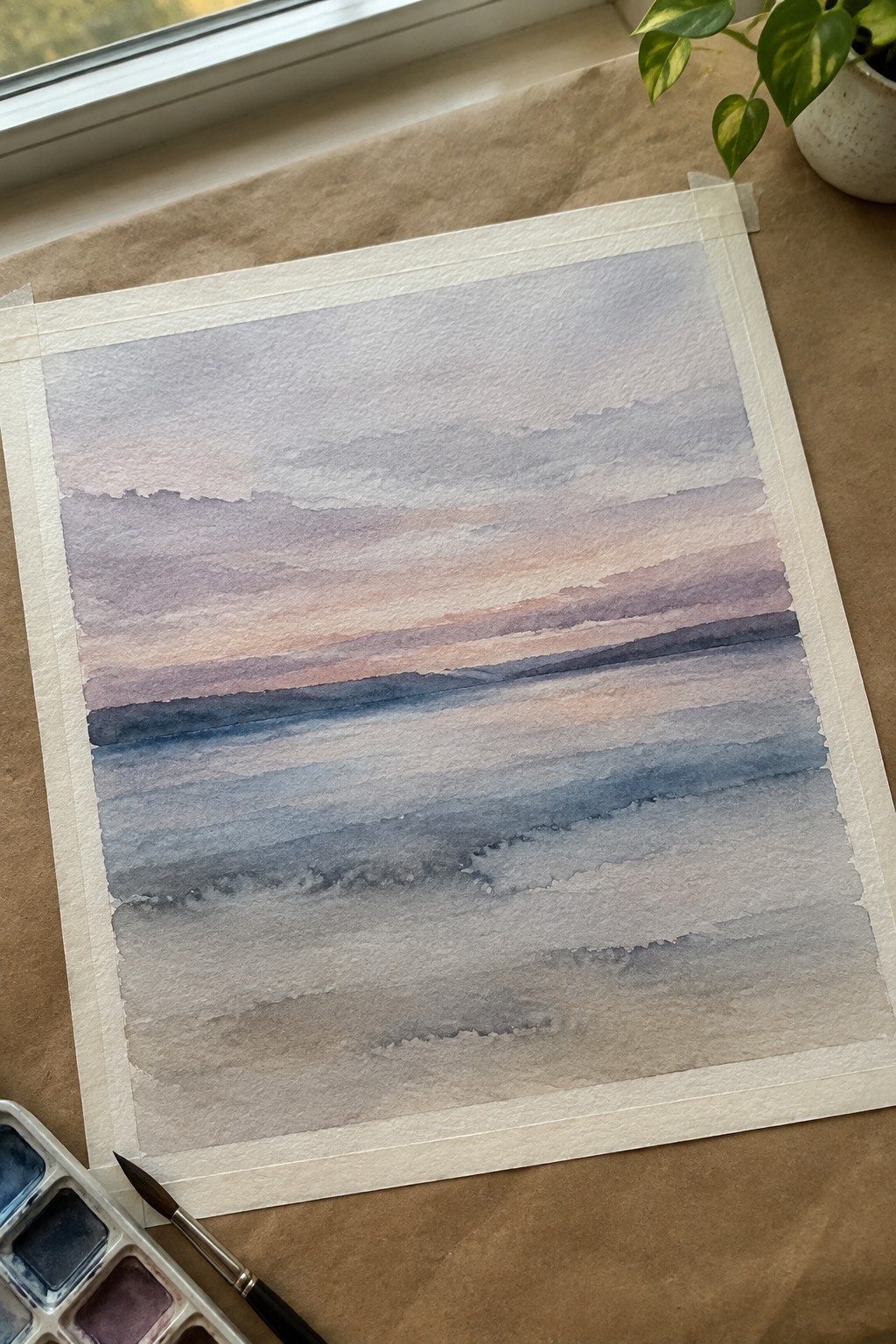 Watercolor painting with horizontal bands of blended purple, pink, blue, and gray tones taped to brown paper, with a palette and brush visible at the bottom left.
