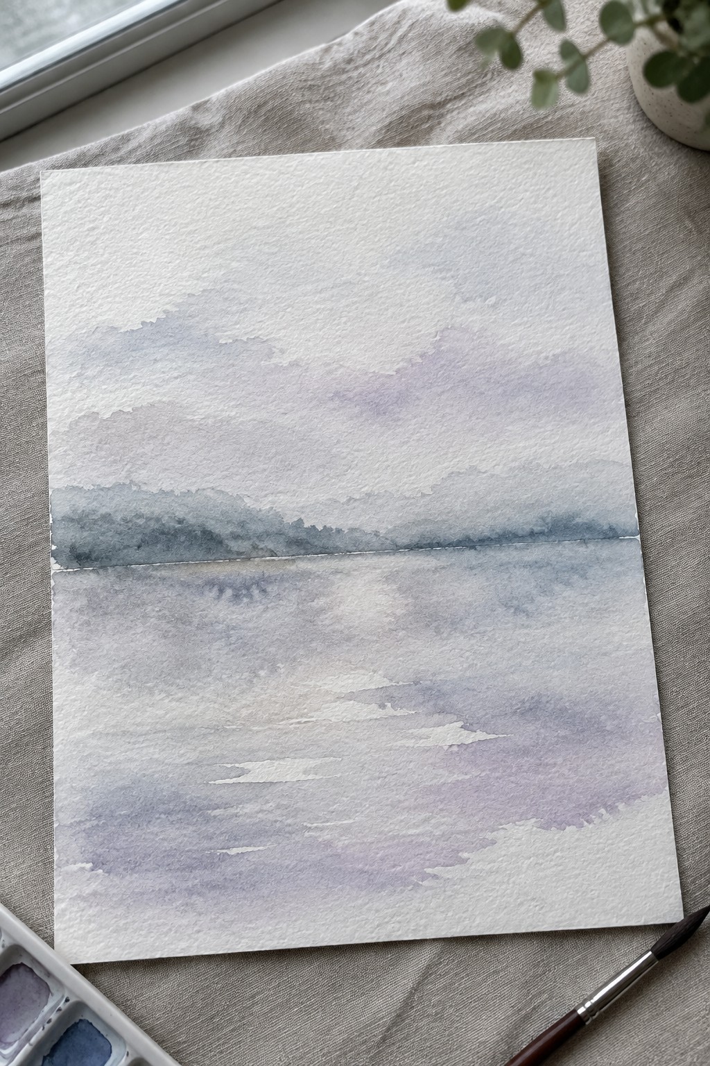 Watercolor landscape in soft blue and purple hues on linen with palette and brush nearby.