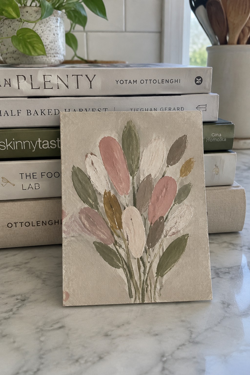 Small abstract floral painting in muted pinks and greens leaning against cookbook stack