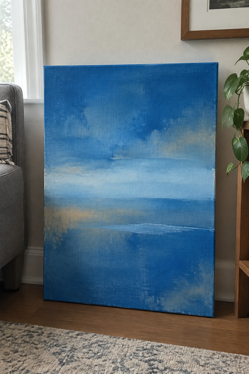 Abstract blue painting on canvas leaning against wall beside gray chair and plant