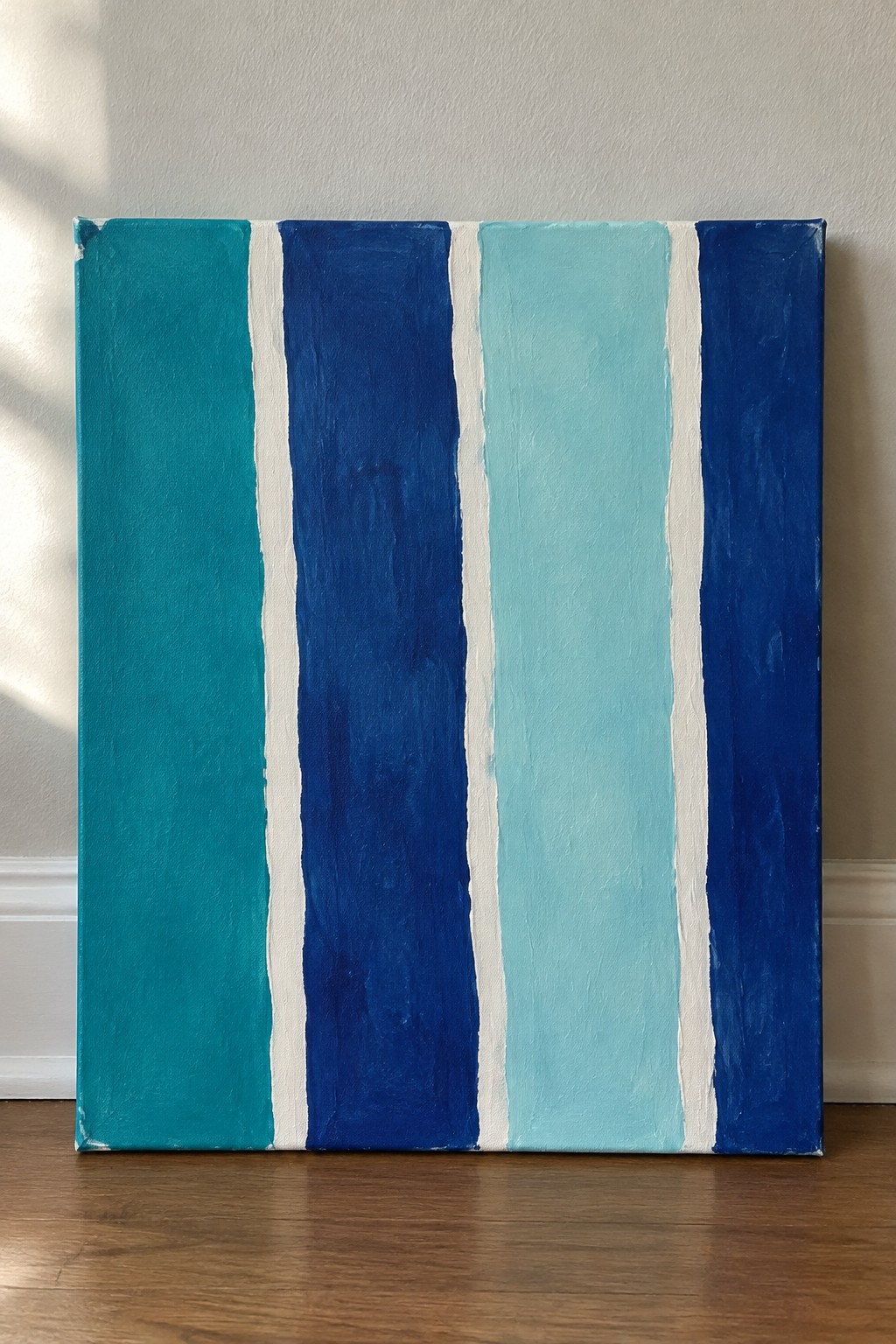 A rectangular canvas with five vertical stripes in teal, dark blue, light cyan, and navy separated by thin white lines, propped against a wall on a wooden floor.