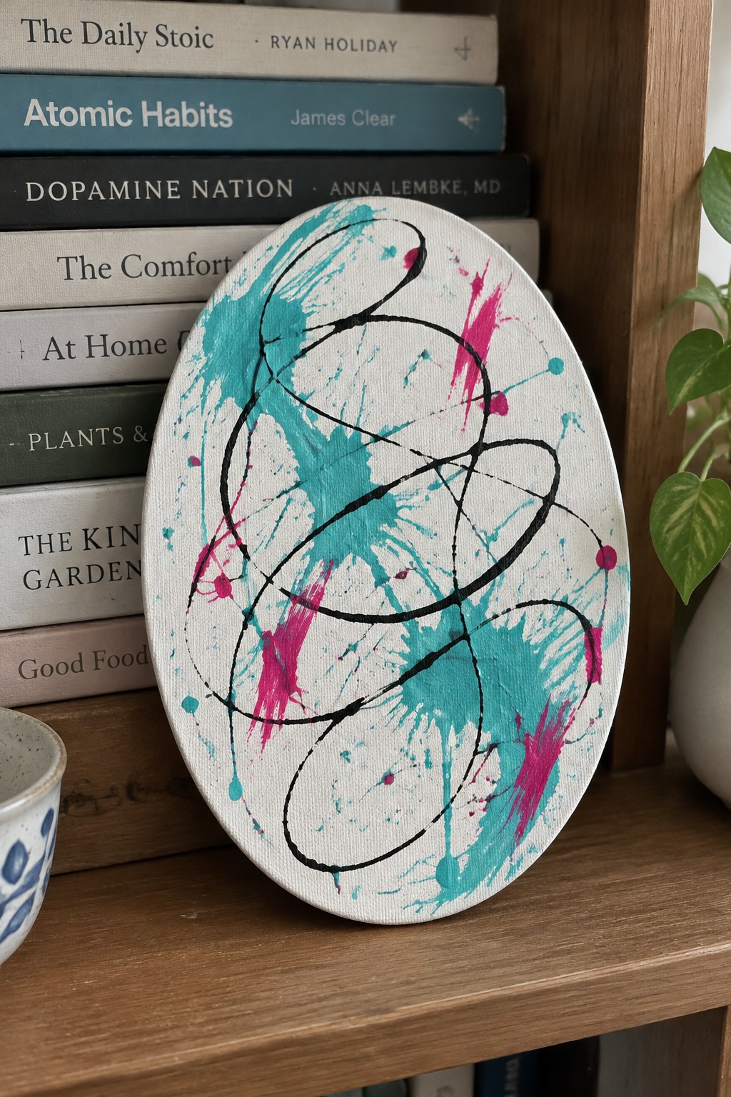 Abstract oval canvas with teal, black, and magenta splatters on bookshelf with books