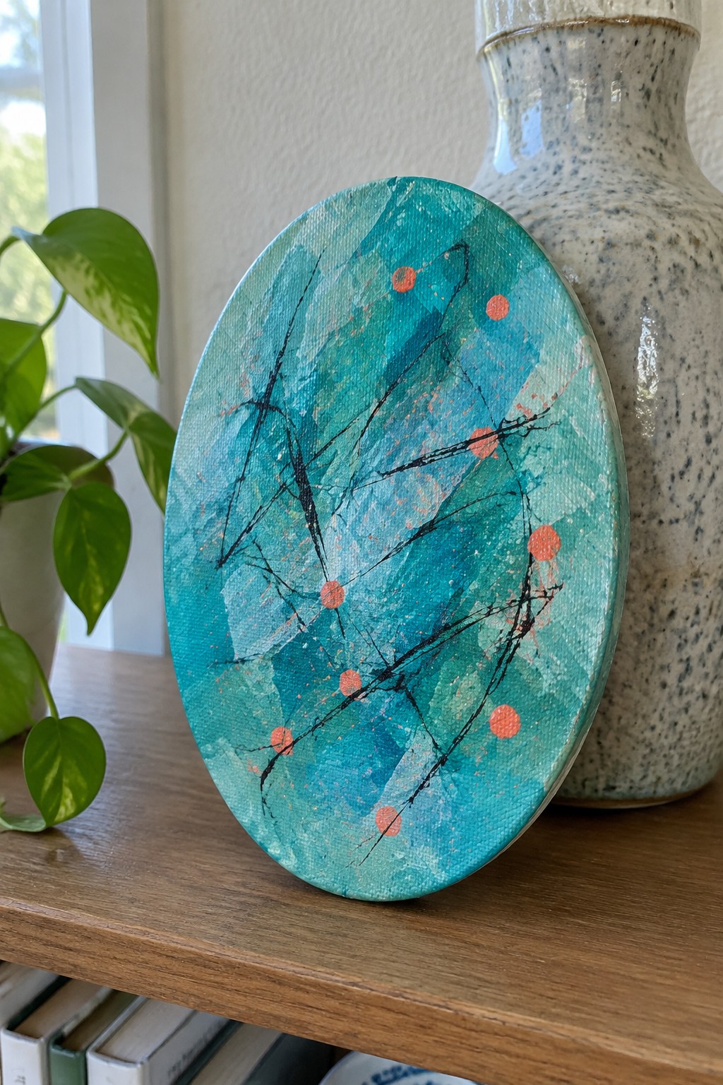 Oval turquoise abstract painting with black lines and orange dots beside ceramic vase on shelf