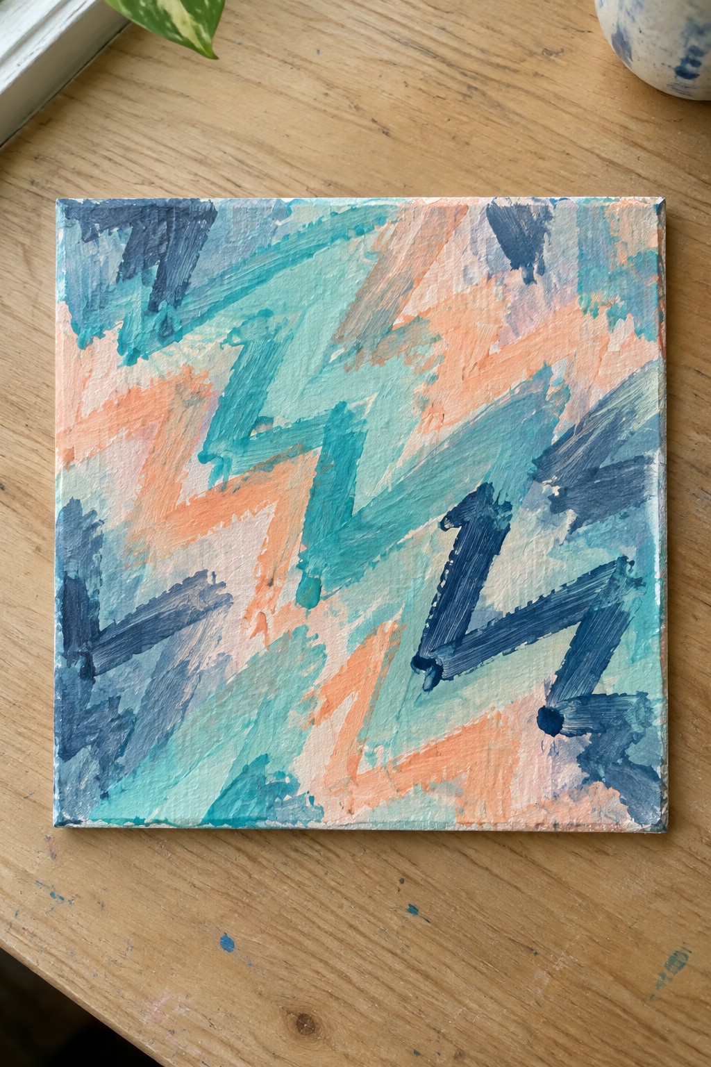 Abstract painting on square canvas with blue, teal, and orange brushstrokes on wooden table