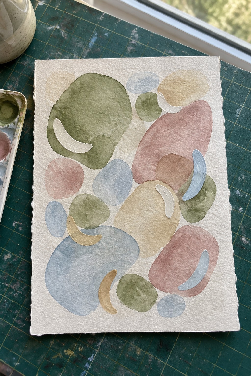 Watercolor painting on textured white paper with overlapping organic shapes in muted green, pink, blue, beige, and gray tones plus a few white highlights.