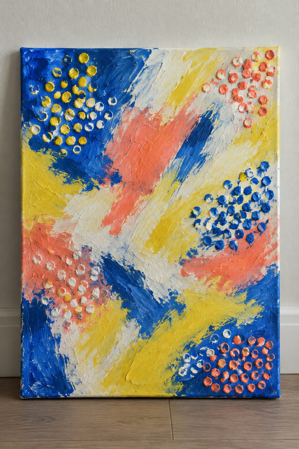 Abstract painting on canvas with bold blue, yellow, orange strokes and clustered paint circles against wall.