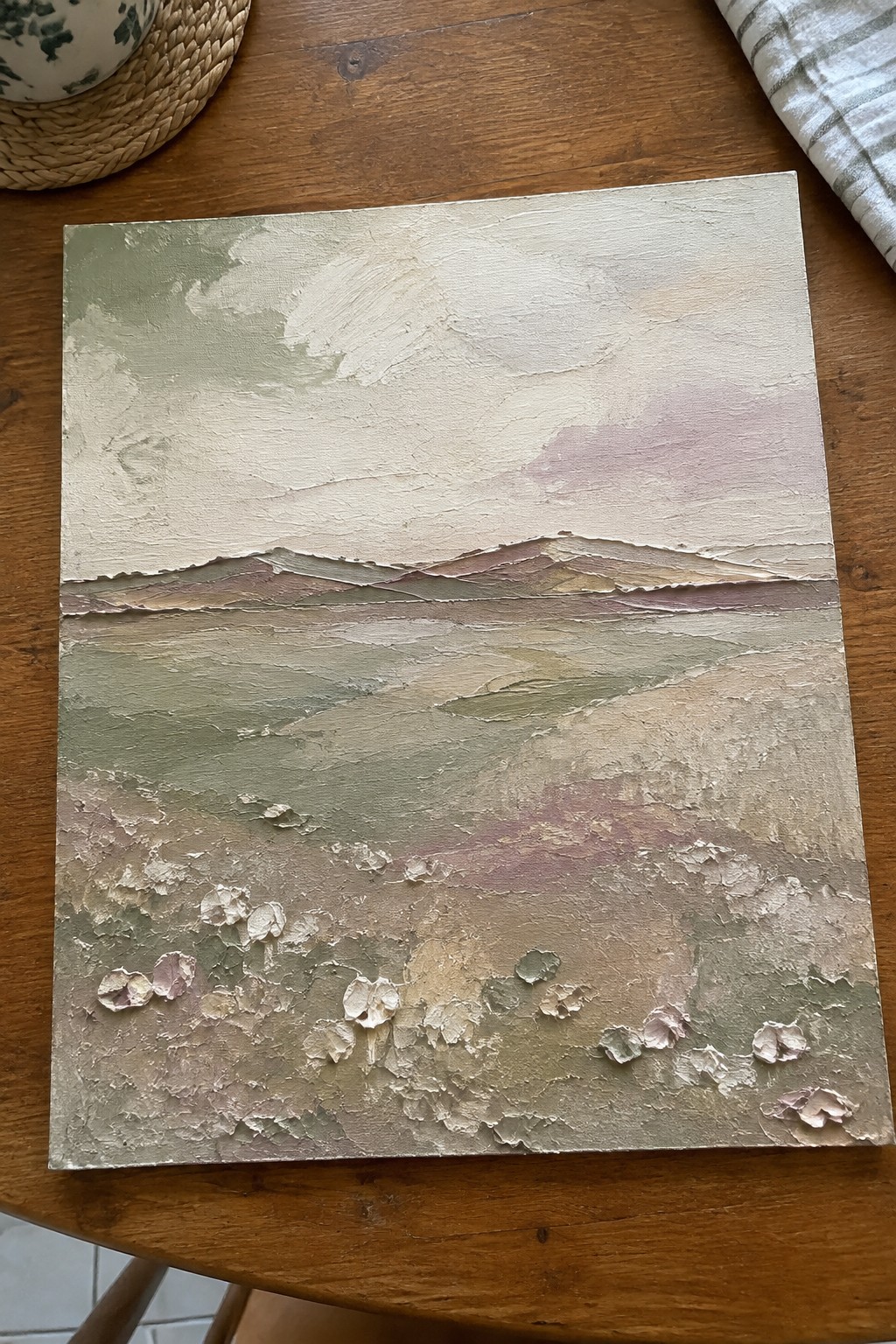 Textured abstract landscape painting in muted greens, pinks, and creams on wooden table