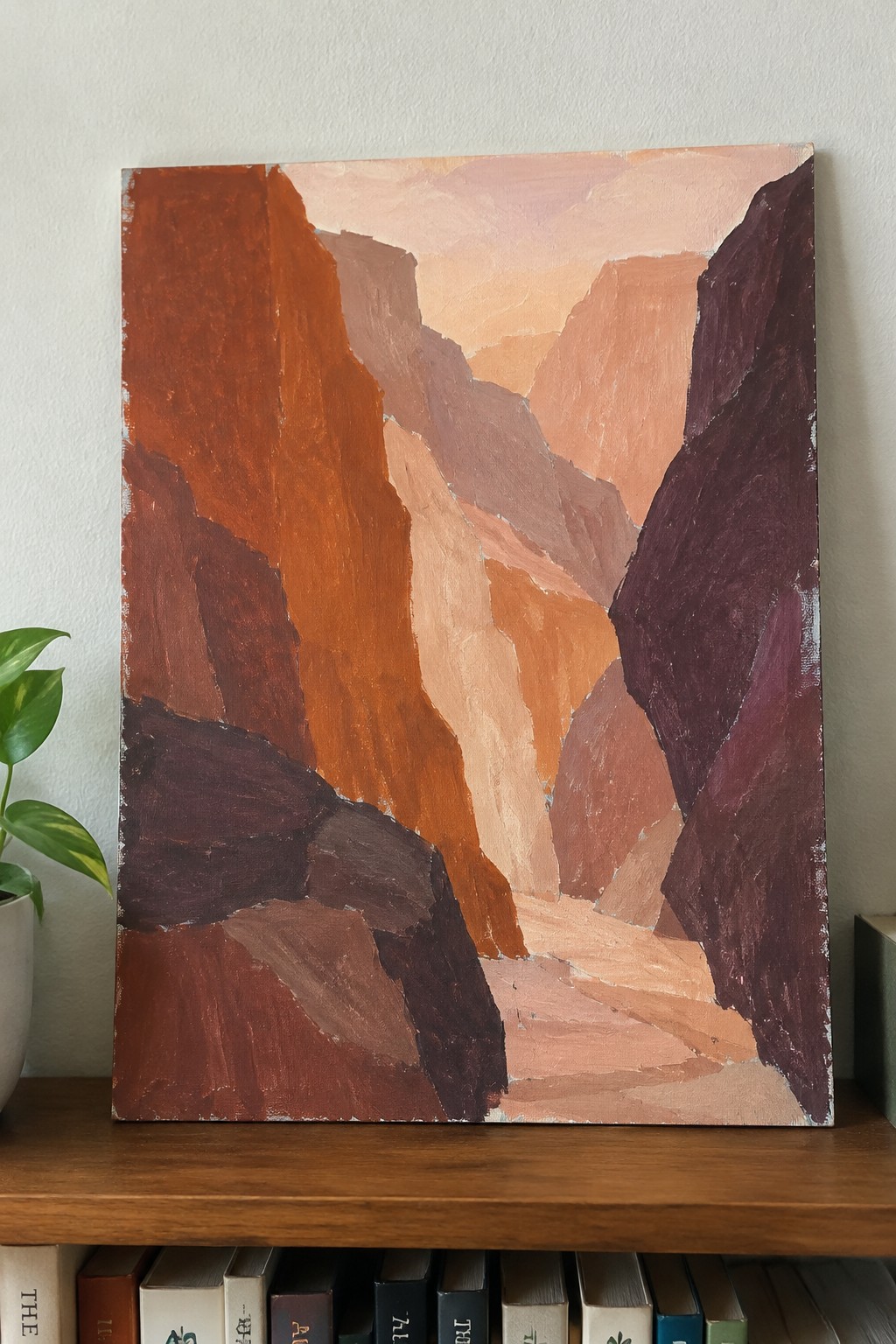 Abstract painting of rocky canyon in warm orange, red and purple tones on wooden shelf above books.
