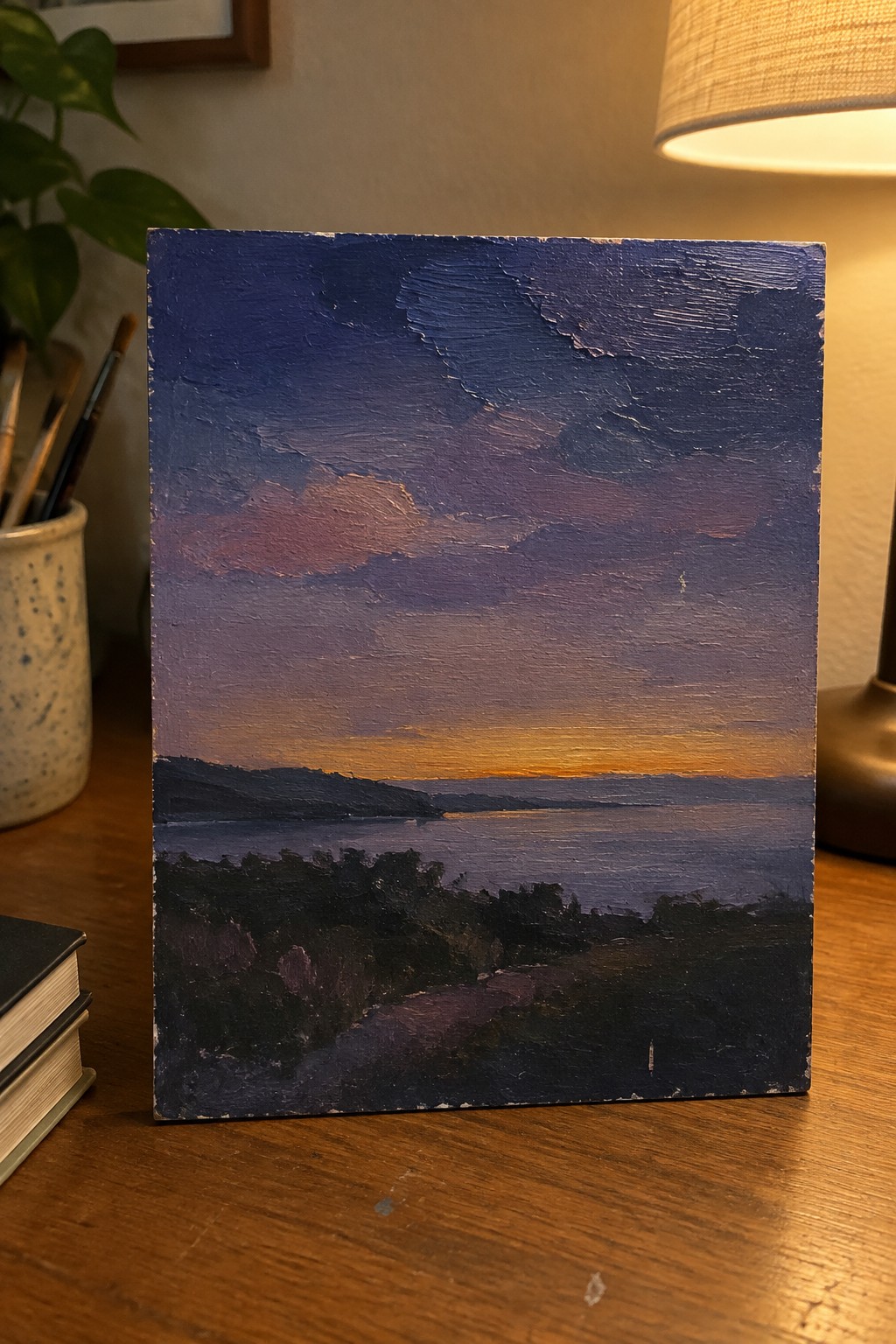 Textured oil painting of purple sunset over water on wooden desk beside lamp
