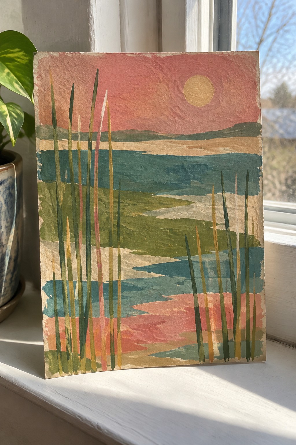 Colorful abstract landscape painting with pink sky, sun, layered hills, and reeds on windowsill.
