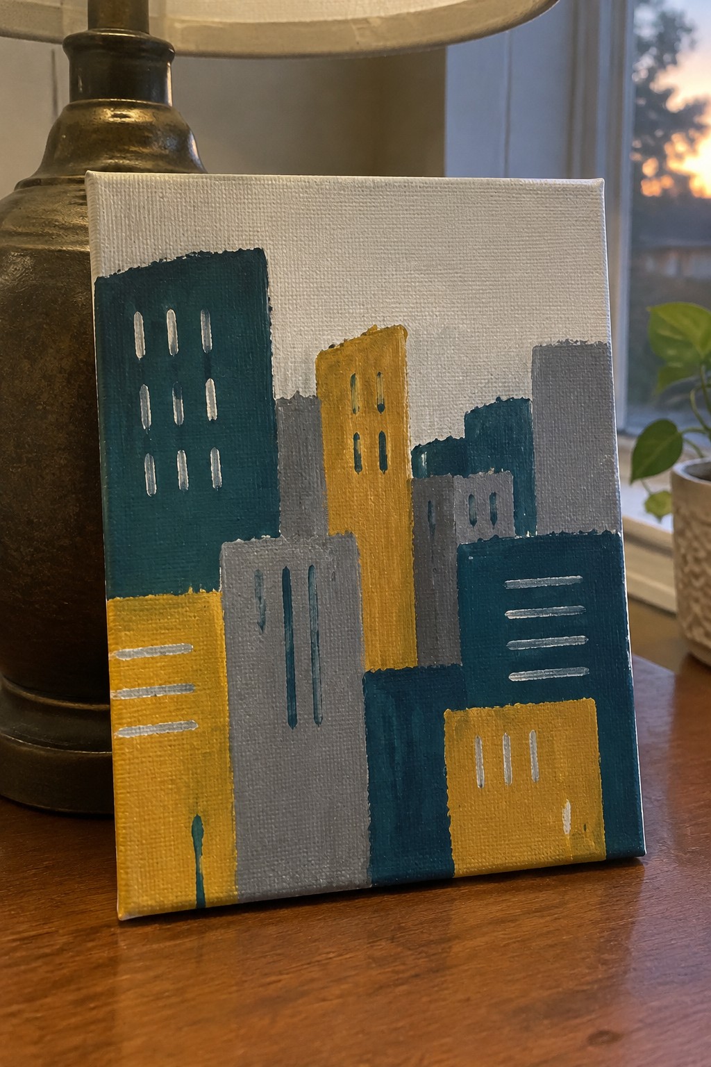 Abstract painting of teal yellow gray city buildings on table beside brass lamp