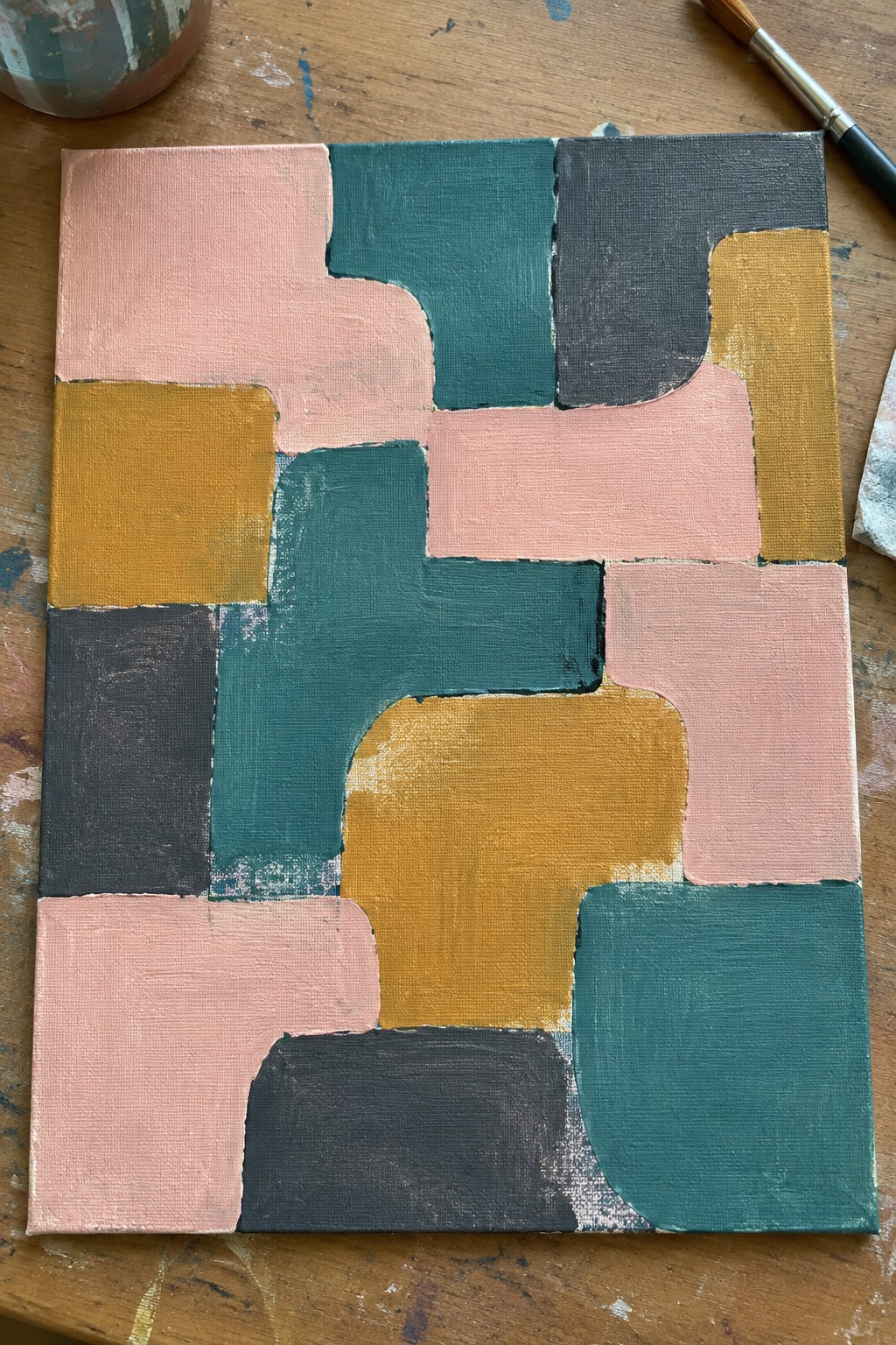 Abstract geometric painting with pink, teal, yellow, and gray shapes on wooden table.