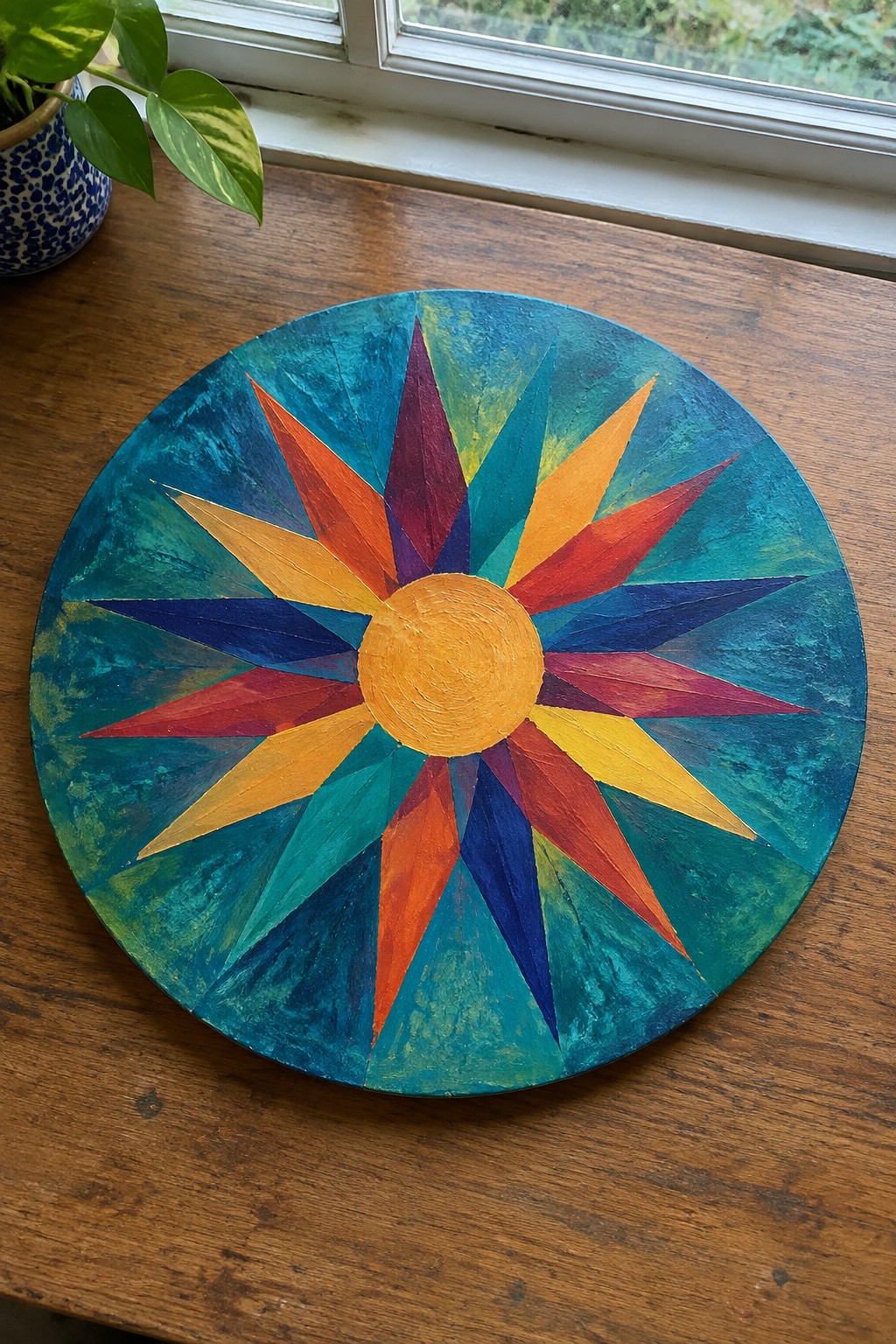 Vibrant multicolored sunburst on round teal canvas atop wooden table with plant nearby